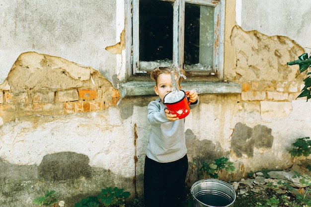 beautiful boy with a white hair red mug and a bucket on the background of the old hut