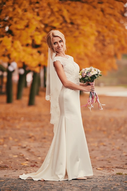 Beautiful bride in luxury white long dress with wedding bouquet in autumn park on wedding day