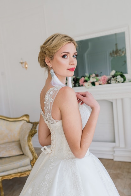 Beautiful bride posing in wedding dress in a white photo studio, mirror and sofa.