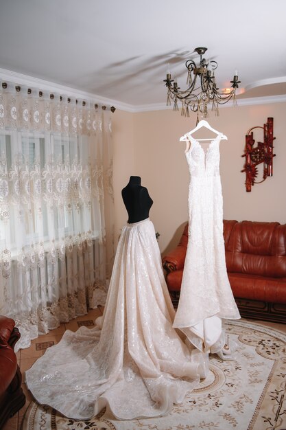 Beautiful bride's white wedding dress hangs near the bed in a hotel room with flowers at the bottom.