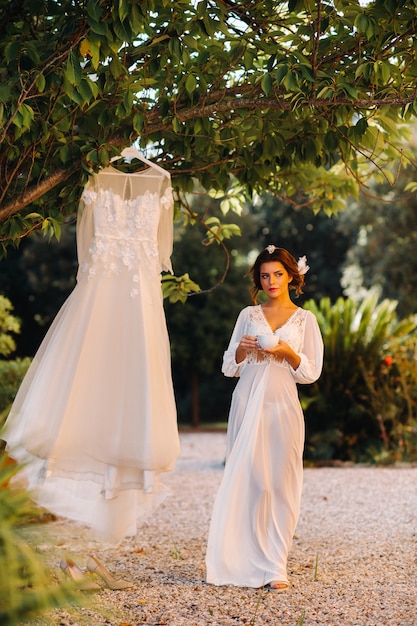 A beautiful bride stands next to a wedding dress with a Cup of tea in a boudoir outfit next to a Villa in Italy.morning of the bride in Tuscany.