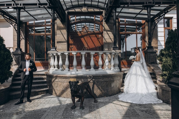 Photo beautiful bride stands in a wonderful dress with a bouquet in her hands near a large gray house