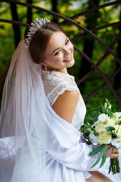 Beautiful bride in veil and white coat sits on a blanket with a wedding bouquet in her hands