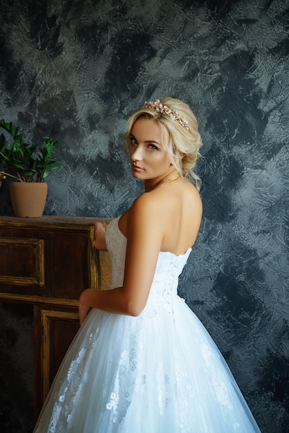 A beautiful bride in a very beautiful long dress with a train stands by the window.