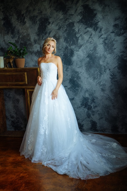 A beautiful bride in a very beautiful long dress with a train stands by the window.