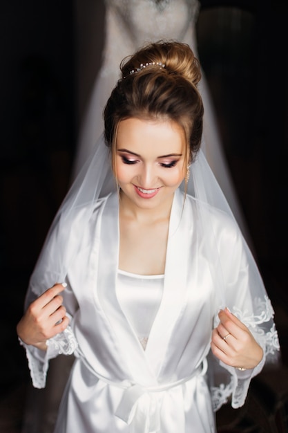 Beautiful bride in white bathrobe with make up and bridal veil closed eyes and smile.