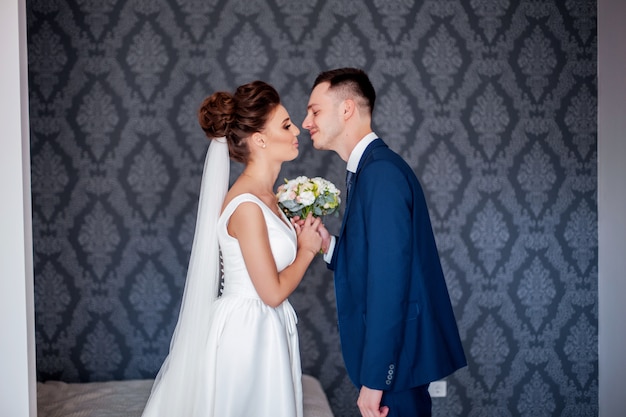 Beautiful bride in white dress with bouquet of brides and handsome groom in blue suit standing at home at home