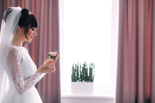 Beautiful bride in a white dress with a veil holds a flower bud in her hands waiting for the groom in the room near the window