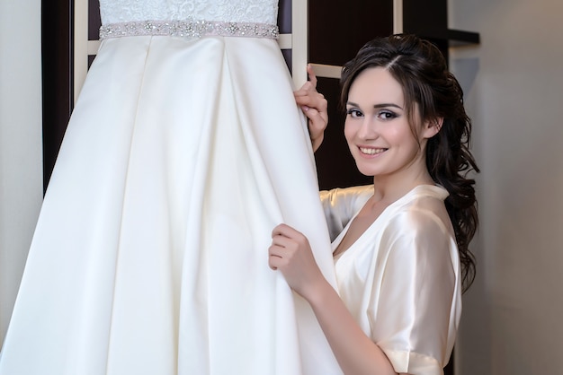 Beautiful bride in white negligee next to her wedding dress smiling