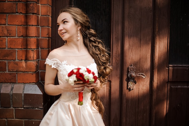 Beautiful bride in white wedding dress with a wedding bouquet standing near the wooden door of old building