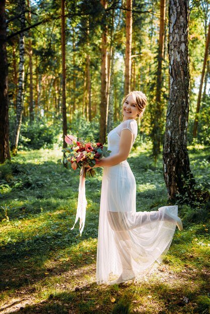 Beautiful bride with a bouquet in her hand posing on her wedding day among the green trees