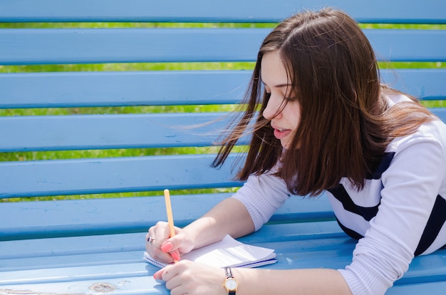 Beautiful brinette girl sitting on a bench, dreaming and writing her plans in a notebook, relaxing in a park on a sunny spring day