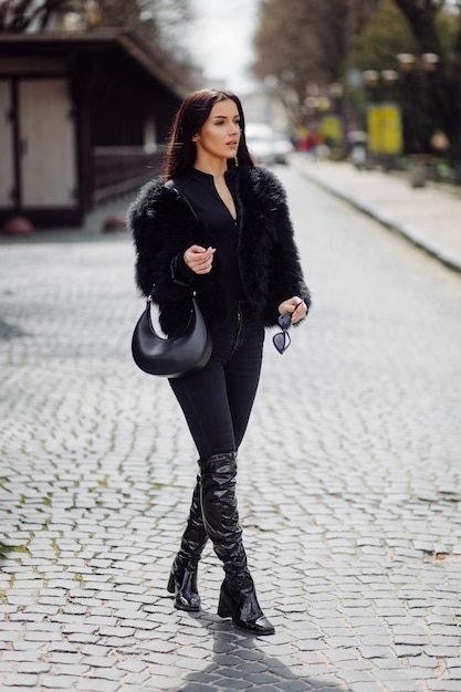 Beautiful brown-haired stylish girl in black dress outdoor. Young attractive elegant woman portrait with long hair on springtime in city streets.