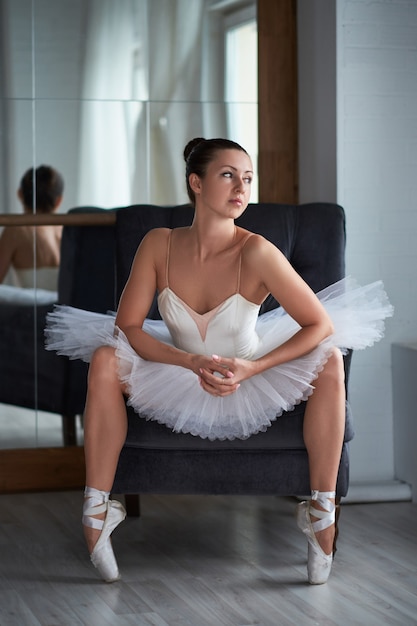 Beautiful brunette ballerina sitting on vintage armchair and adjusting her pointer shoe. Copyspace