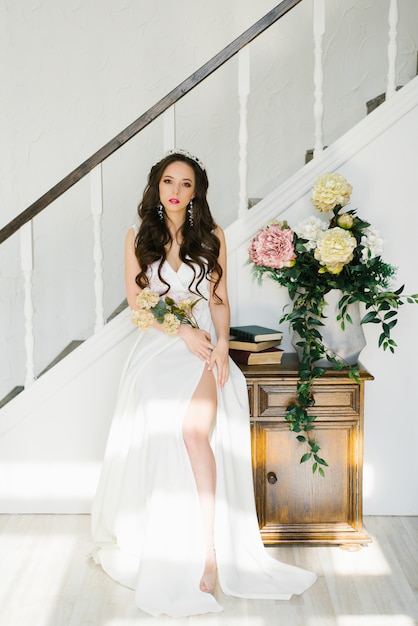 Beautiful brunette bride in a white wedding dress with a slit on the leg in a classic interior. She holds in her hands the flowers