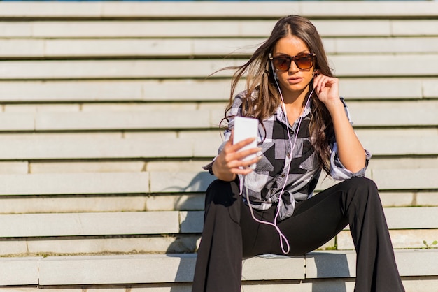 Beautiful brunette caucasian girl listening to the music with headphones on stairs in the city