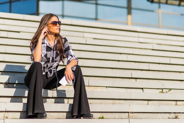 Beautiful brunette caucasian girl listening to the music with headphones on stairs in the city