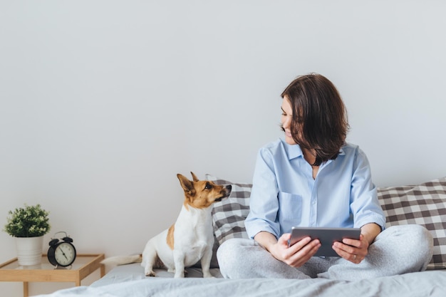 Beautiful brunette female in pyjamas sits on bed in bedroom with her jack russell terrier dog holds tablet computer enjoys morning weekend watches video online has good relationships with pet