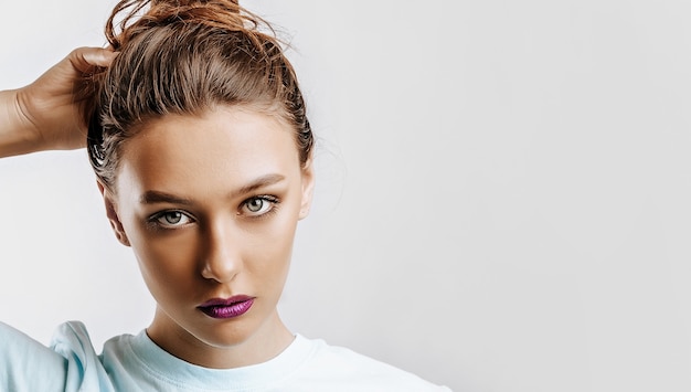 A beautiful brunette girl holds her hair and makes her own hairstyle. Young model with long hair, fashion makeup with purple lips on an gray background. The woman seriously looking at the camera.