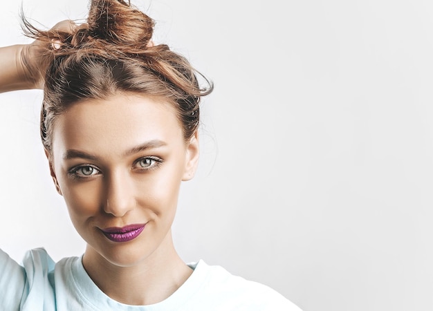 A beautiful brunette girl holds her hair and makes her own hairstyle Young model with long hair fashion makeup with purple lips on an gray background The woman smiles and looking at the camera