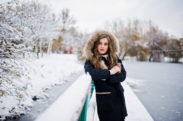 Beautiful brunette girl in winter warm clothing. Model on winter jacket against frozen lake at park.