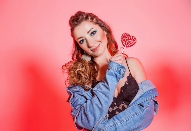 Beautiful brunette girl with blue makeup, pigtails and a sleeveless denim vest posing on a red background with a heart-shaped lollipop. Horizontal photo