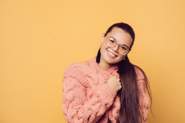 Beautiful brunette in glasses with long hair dressed in pink knitted sweater with a kind smile, looks with tender, over yellow background with copy space. Warm emotions. Being in love.