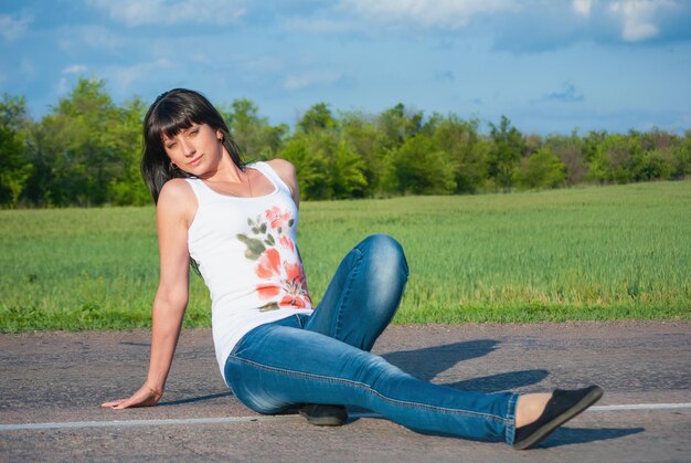 A beautiful brunette in jeans with big breasts sits against the blue sky in a white