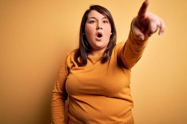 Beautiful brunette plus size woman wearing casual sweater over isolated yellow background Pointing with finger surprised ahead open mouth amazed expression something on the front