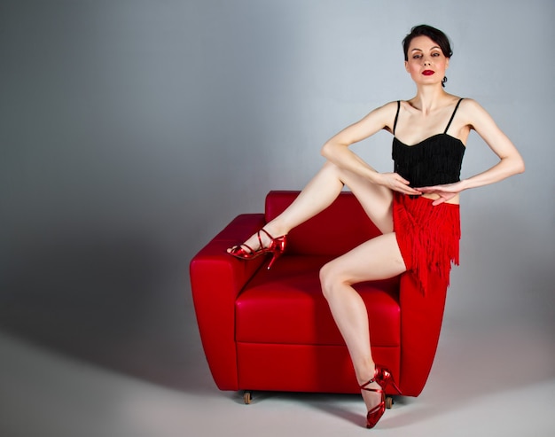 a beautiful brunette in red shoes sits in a red armchair on a gray background.