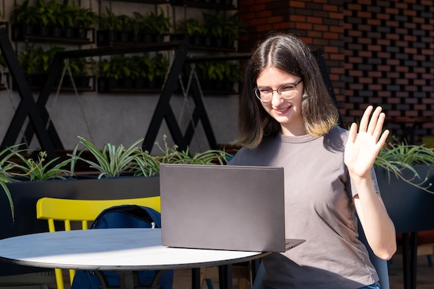 A beautiful brunette student with glasses uses a laptop in a city cafe chatting with friends via video chat greeting