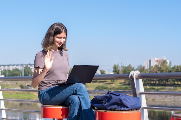 A beautiful brunette student with glasses uses a laptop on the street chatting with friends via video chat greeting