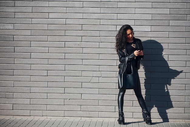 Beautiful brunette with curly hair and in black clothes outdoors near wall