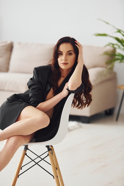 Beautiful brunette with curly hair in black luxury clothes sits on chair at home at daytime