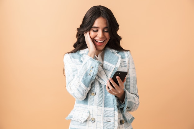 Beautiful brunette woman dressed in plaid jacket standing isolated over beige, holding mobile phone