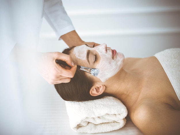 Beautiful brunette woman enjoying applying cosmetic mask with closed eyes comfortable and blissful. Relaxing treatment in medicine and spa center concepts.