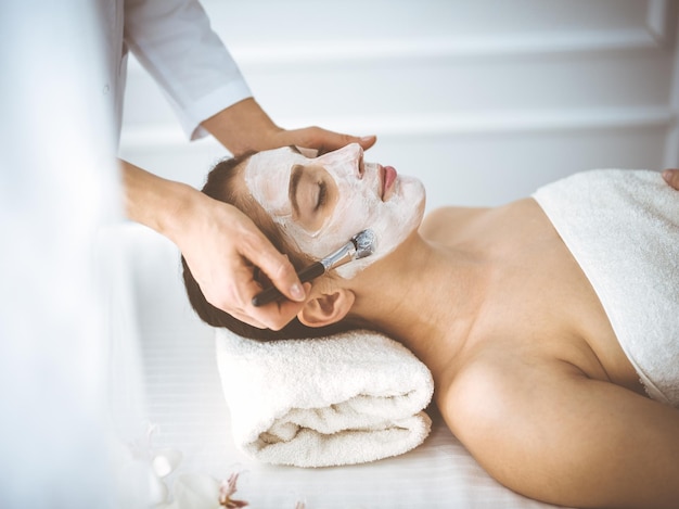 Beautiful brunette woman enjoying applying cosmetic mask with closed eyes comfortable and blissful. Relaxing treatment in medicine and spa center concepts.