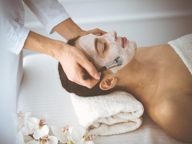 Beautiful brunette woman enjoying applying cosmetic mask with closed eyes comfortable and blissful. Relaxing treatment in medicine and spa center concepts.