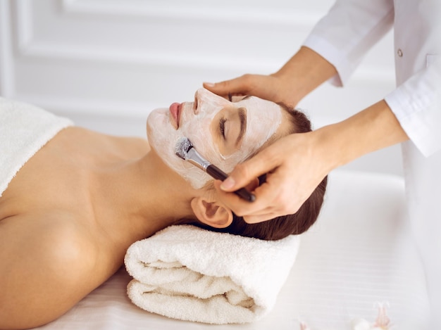 Beautiful brunette woman enjoying applying cosmetic mask with closed eyes. Relaxing treatment in medicine and spa center concepts.