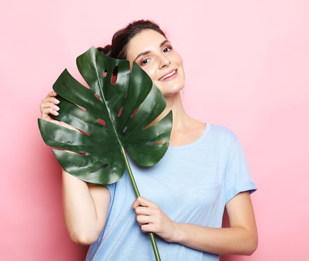 Beautiful brunette woman holding a leaf of a large tropical flower and smiling over pink background