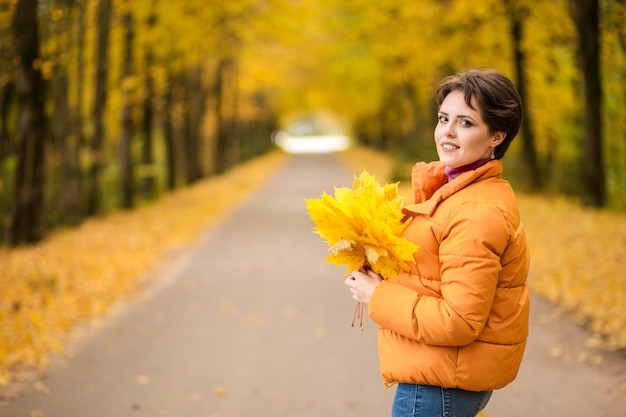 Beautiful brunette woman posing in a yellow jacket in an autumn park with a bouquet of autumn leaves and looking at the camera