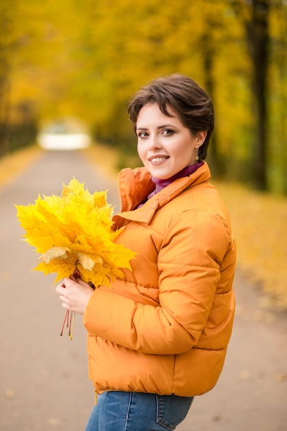 Beautiful brunette woman posing in a yellow jacket in an autumn park with a bouquet of autumn leaves and looking at the camera