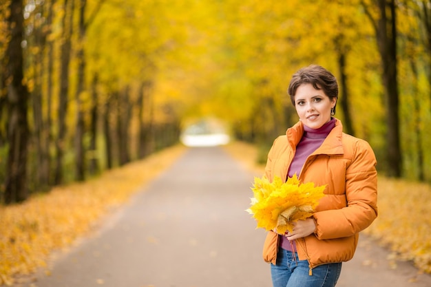 Beautiful brunette woman posing in a yellow jacket in an autumn park with a bouquet of autumn leaves