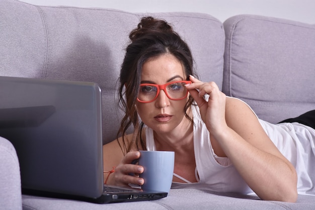 Beautiful brunette woman in red glasses on sofa using laptop