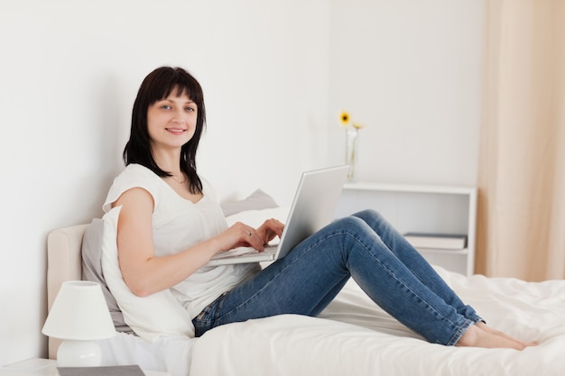 Beautiful brunette woman relaxing with her laptop while sitting on a bed