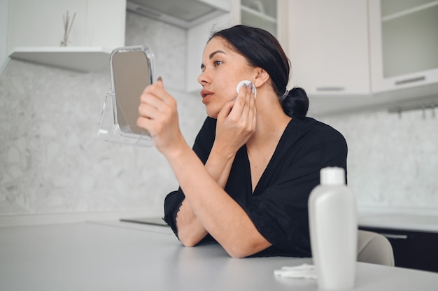 Beautiful brunette woman removing makeup from her face with mirror. Cute beautiful natural beauty girl cleaning face with cotton sponge. Cosmetology and spa, problem skin care, acne treatment concept