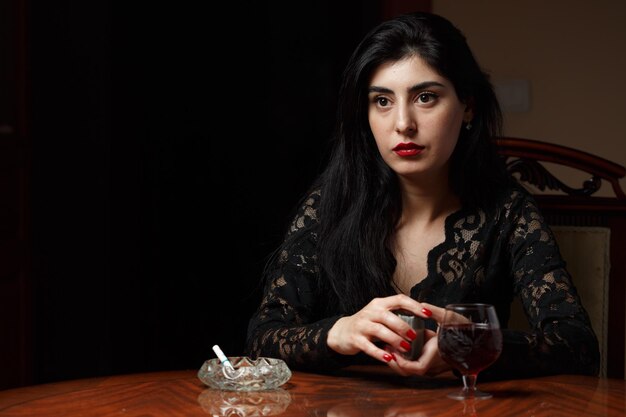 A beautiful brunette woman with scarlet lips in black clothes sits at a table and plays cards for undressing