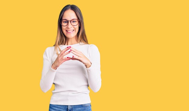 Beautiful brunette young woman wearing casual white sweater and glasses hands together and fingers crossed smiling relaxed and cheerful. success and optimistic