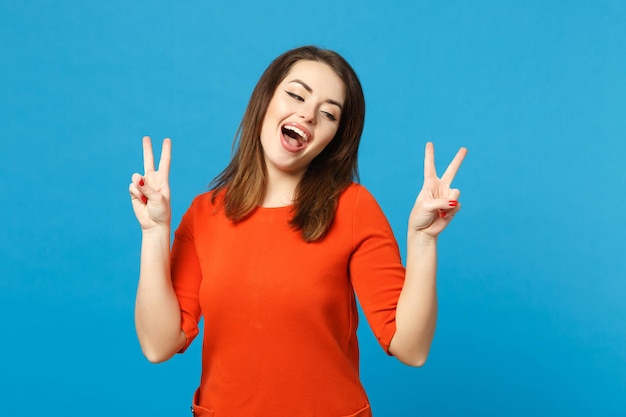 Beautiful brunette young woman wearing red orange dress looking camera showing victory sign isolated over blue wall background, studio portrait. People lifestyle fashion concept. Mock up copy space.