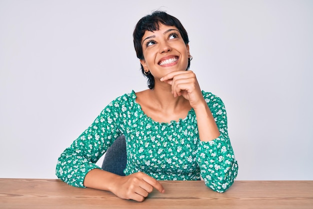 Beautiful brunettte woman wearing casual clothes sitting on the table looking confident at the camera with smile with crossed arms and hand raised on chin thinking positive